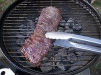 using tongs to grab a piece of flank steak on a charcoal grill