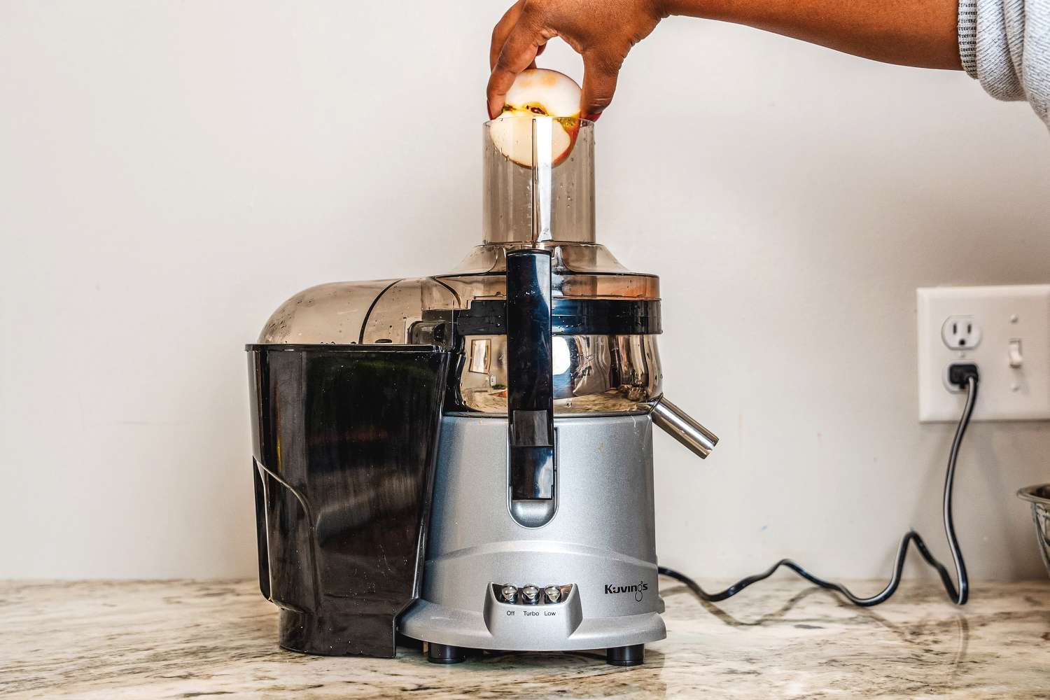 A person places half an apple in a Kuvings centrifugal juicer's chute on a granite counter