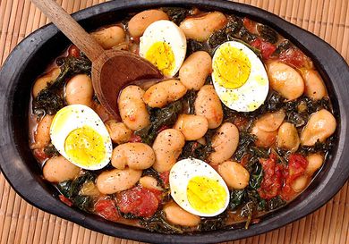 Overhead view of butter beans with kale and eggs, served in a black clay platter.
