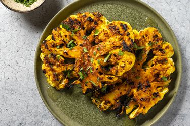 A head of spiced and grilled cauliflower, cut in half lengthwise and overlapping on a green ceramic plate. At the top left edge of the image is a small ceramic bowl holding chopped herbs, which are also sprinkled across the surface of the cauliflower.