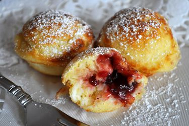 Three ebelskivers on a white cloth, dusted with powdered sugar. One is torn is cut in half, showing the lingonberry jam filling.