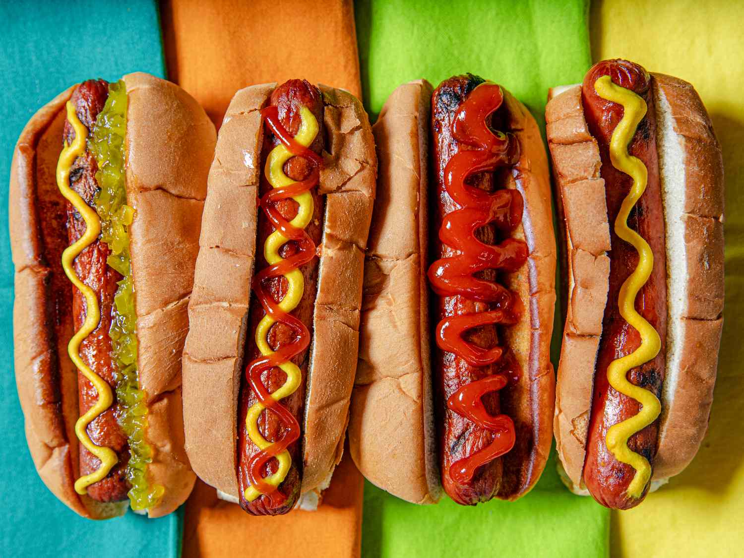 Four hot dogs with various condiments lined up on colorful backgrounds