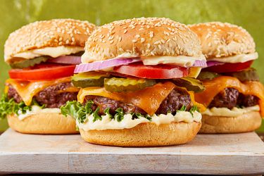 Three cheeseburgers with lettuce, tomato, pickle, onion, and cheddar cheese on a sesame seed bun displayed on a cutting board