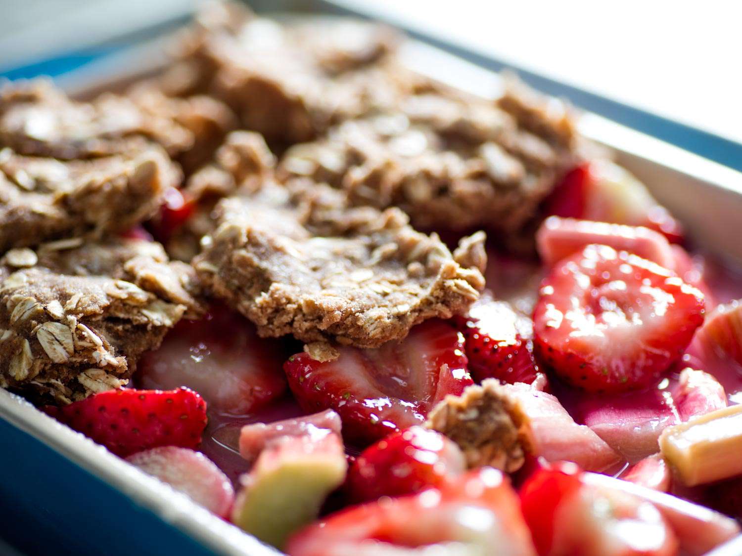Flattened dollops of streusel topping being placed over a filling for strawberry-rhubarb crisp in a blue ceramic baking dish