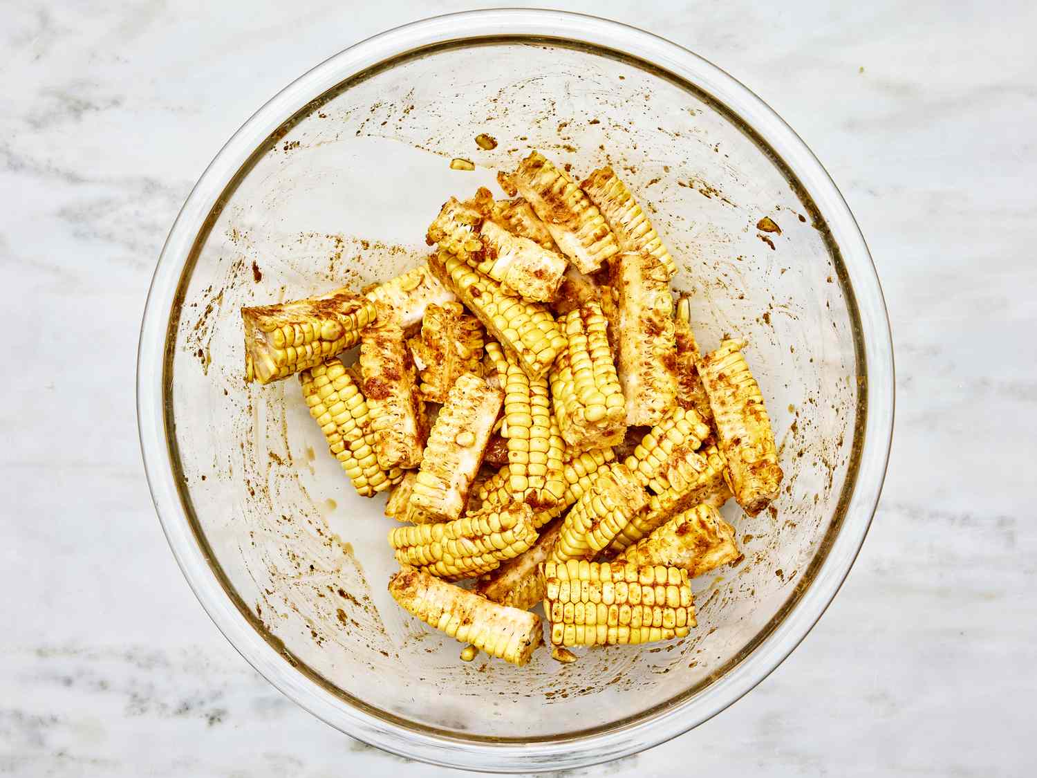 overhead angle of glass bowl on a marble surface. Inside the glass bowl is pieces of corn marinating in spices