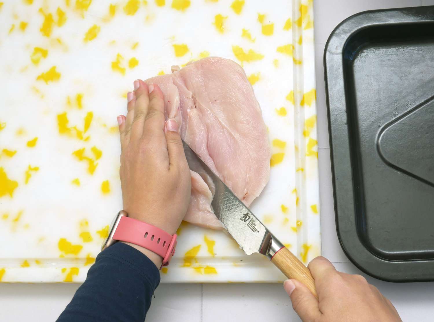 A persons hands cutting a chicken fillet with a knife on a cutting board