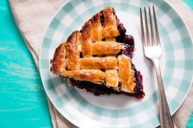 A wedge of herringbone lattice–topped blueberry pie on a plaid-rimmed plate with a fork