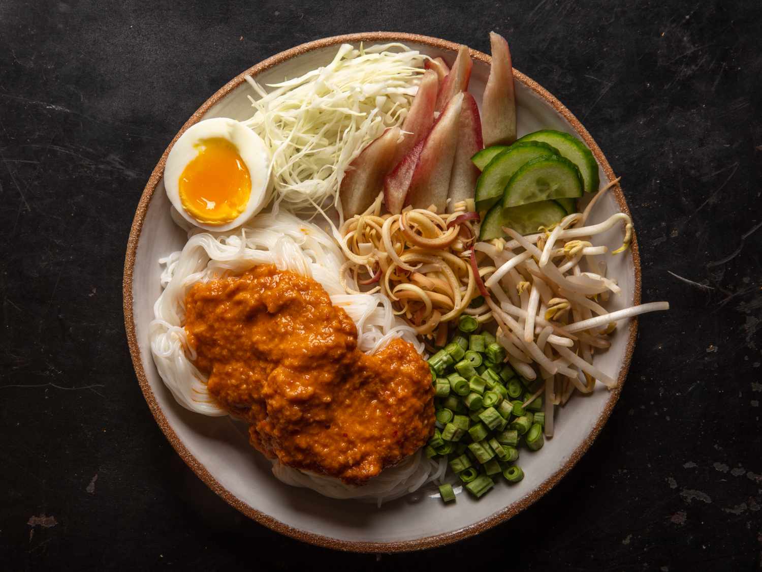 Overhead of a serving plate of noodles dressed with fish curry, with assorted vegetable garnishes and a soft-cooked egg on the side.