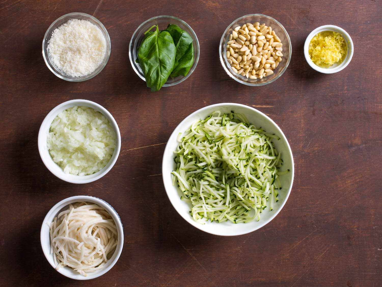 Overhead shot of bowls of ingredients for making zucchini-Parmesan latkes: shredded zucchini, shredded potato, chopped onion, grated Parmesan, basil leaves, pine nuts, and lemon zest