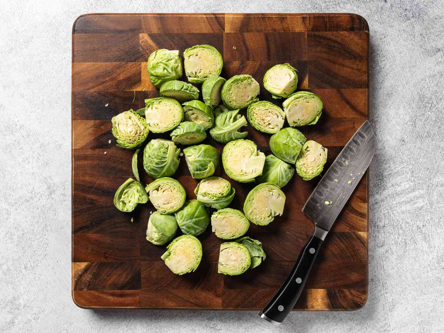 A cutting board holding brussels sprouts which have been trimmed and cut in half. A knife rests on the bottom right hand corner of the cutting board.