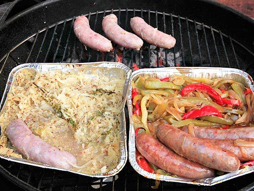 Two aluminum trays of sausages (one with sauerkraut and mustard, one with peppers and onions) on the grill