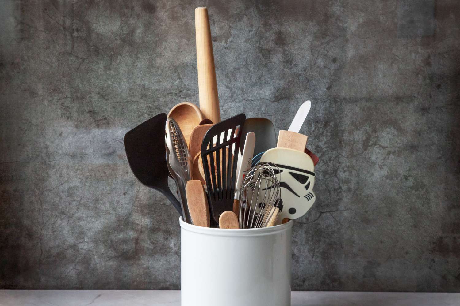 A white ceramic utensil crock filled with various kitchen tools, including a wooden rolling pin.
