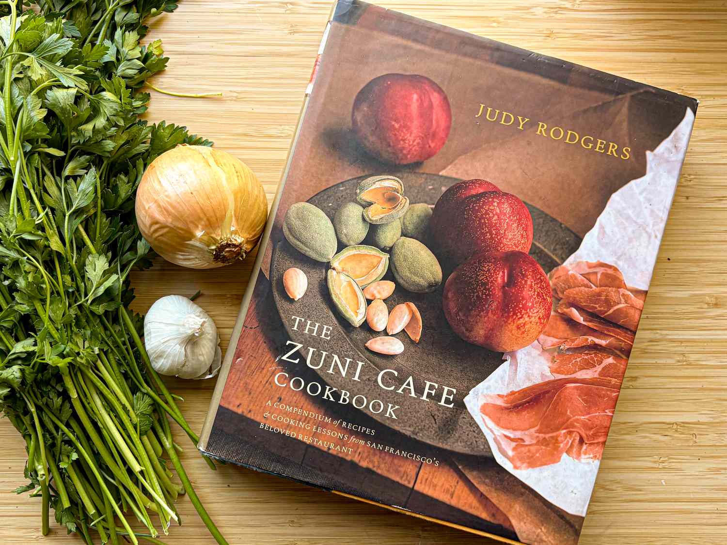 A cookbook titled The Zuni Cafe Cookbook by Judy Rodgers next to parsley an onion and garlic on a wooden surface