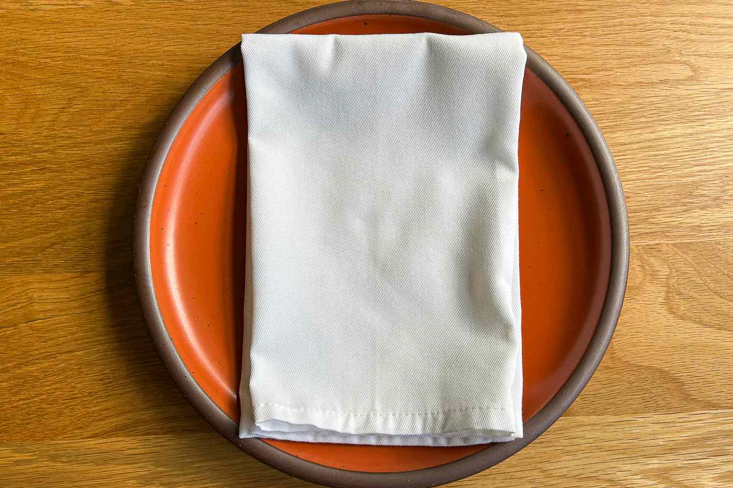 A white napkin neatly folded on an orange plate set on a wooden table