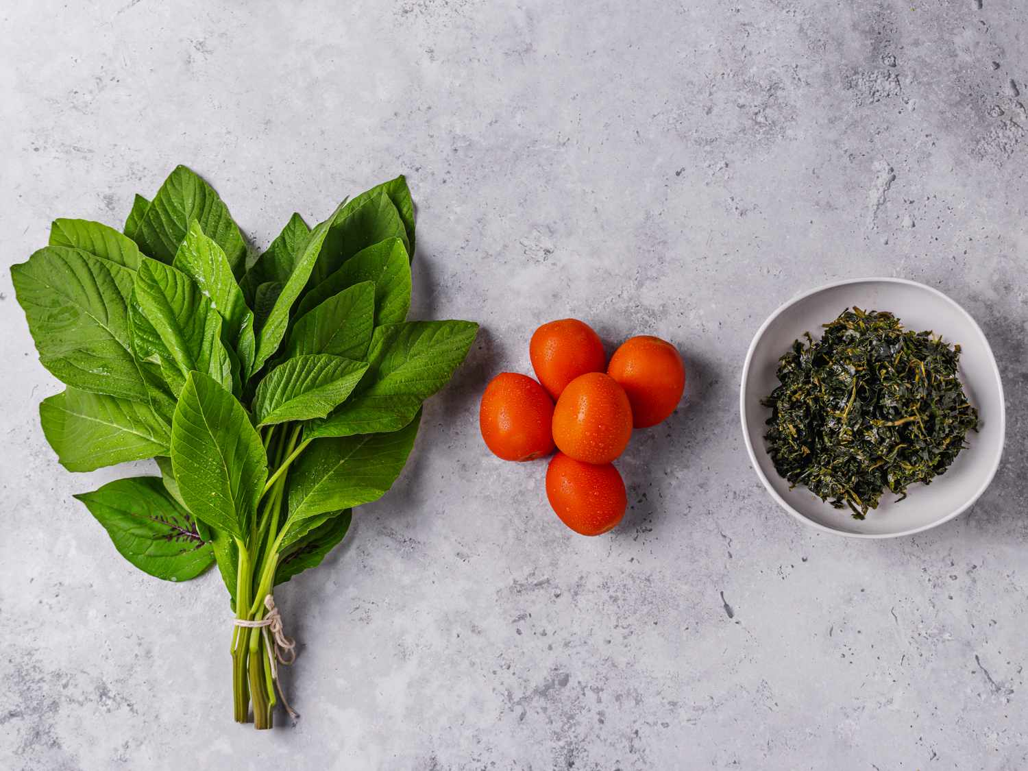 Top down view of amaranth, tomatoes, and dried pumpkin greens in a small bowl arranged in a straight line on a grey marbled backdrop