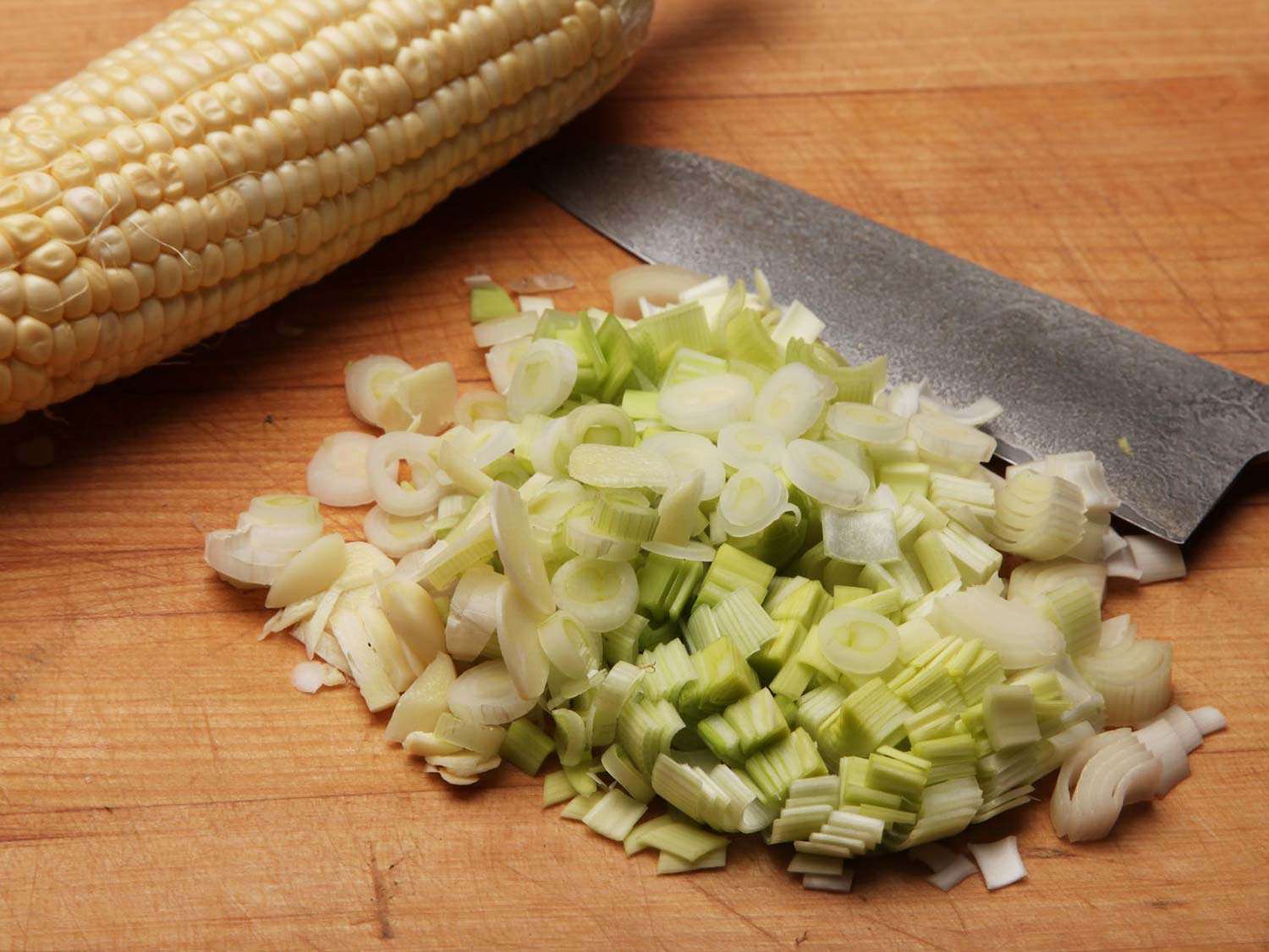 A whole corn cob, a chef's knife, and pile of chopped leeks on a cutting board.