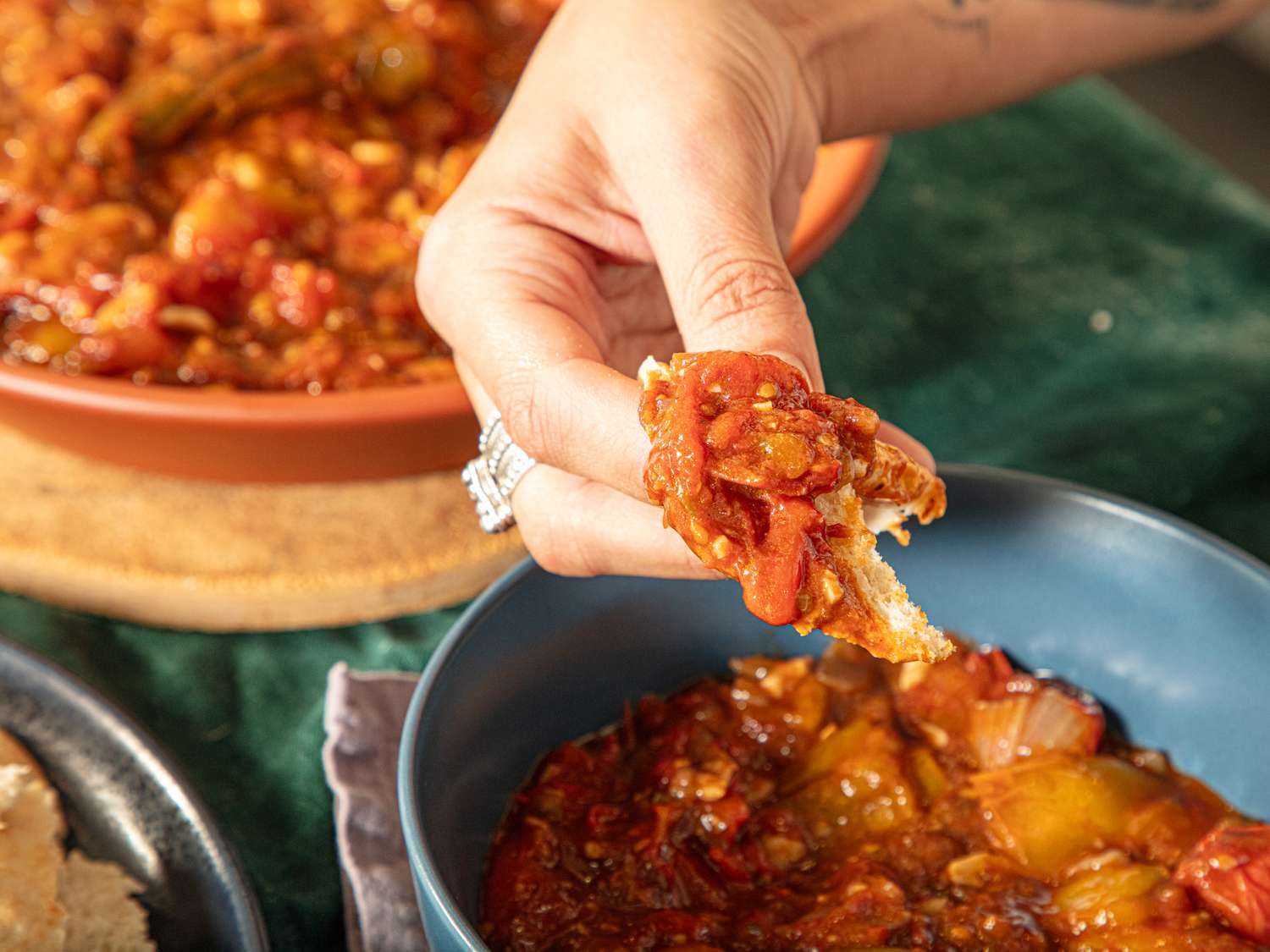 A hand dipping bread into a bowl of a cooked tomatobased dish another dish in the background
