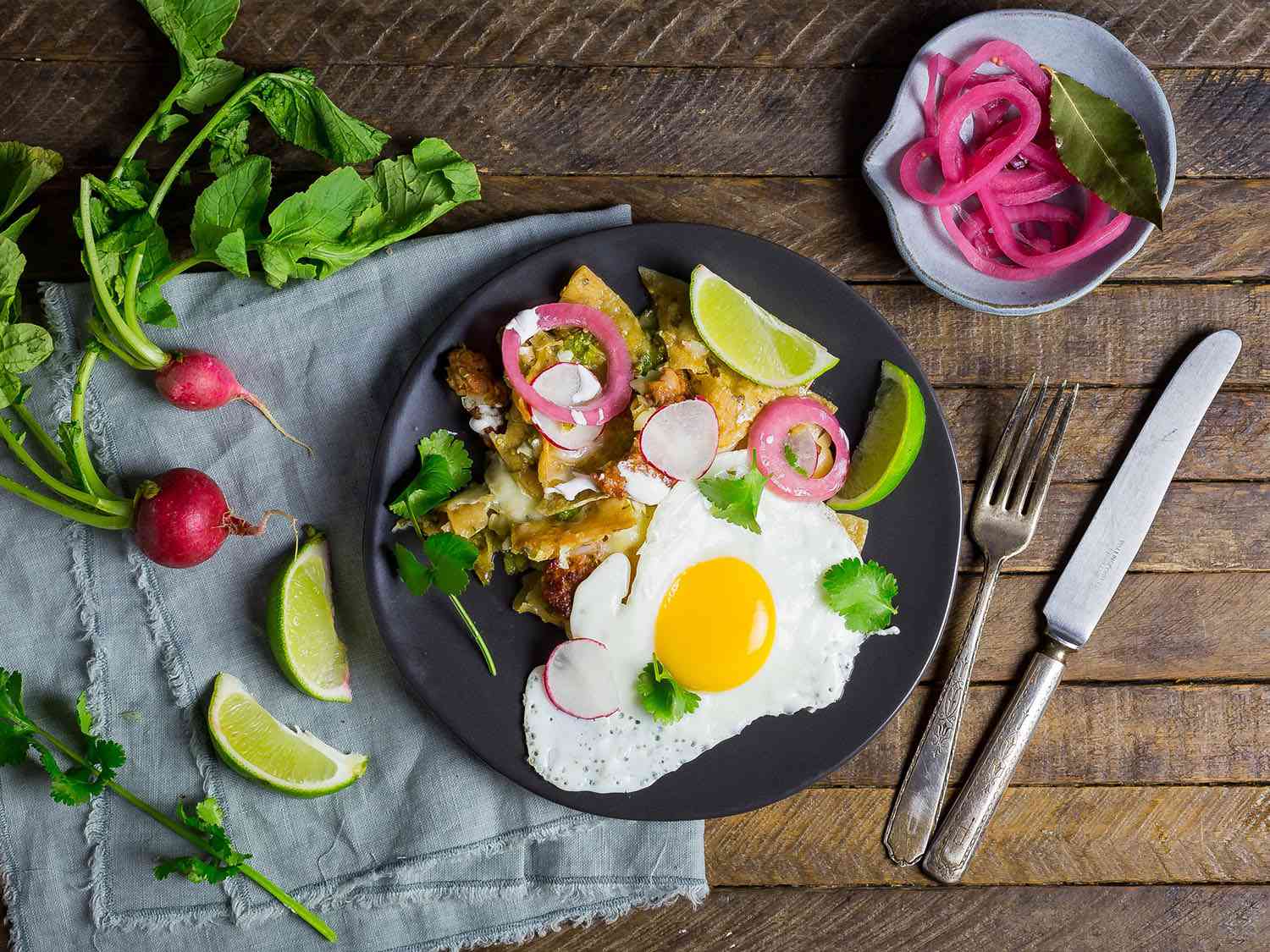 Overhead shot of a plate of homemade tortilla chips covered with salsa verde, topped with sunny-side-up egg, cilantro, and pickled red onion, garnished with lime wedges.