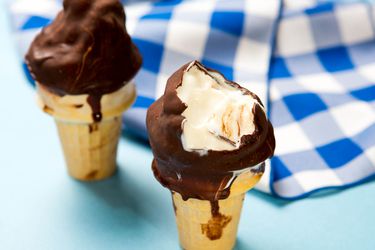 Two homemade chocolate-dipped soft-serve vanilla bean ice cream cones against a blue gingham napkin, a bite taken out of one.