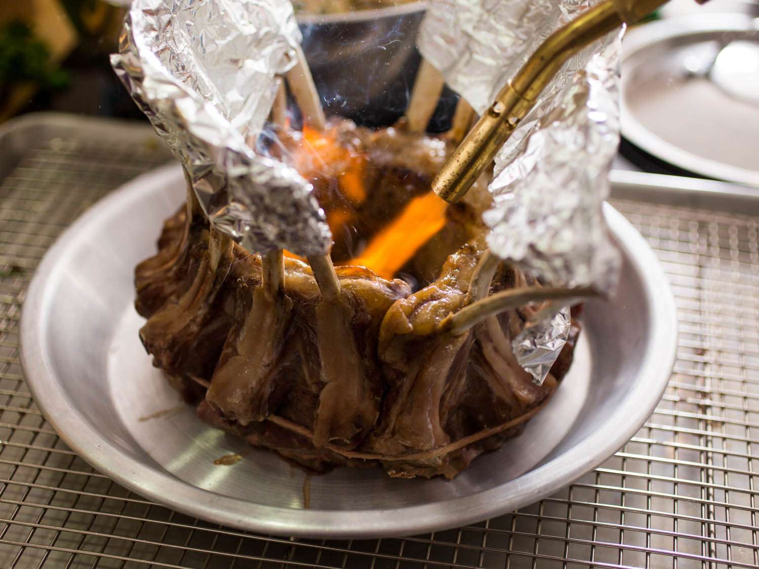 A cooked crown roast of lamb sitting on a plate on a rimmed baking sheet, bones covered in foil, with a blowtorch searing the inner fat cap.