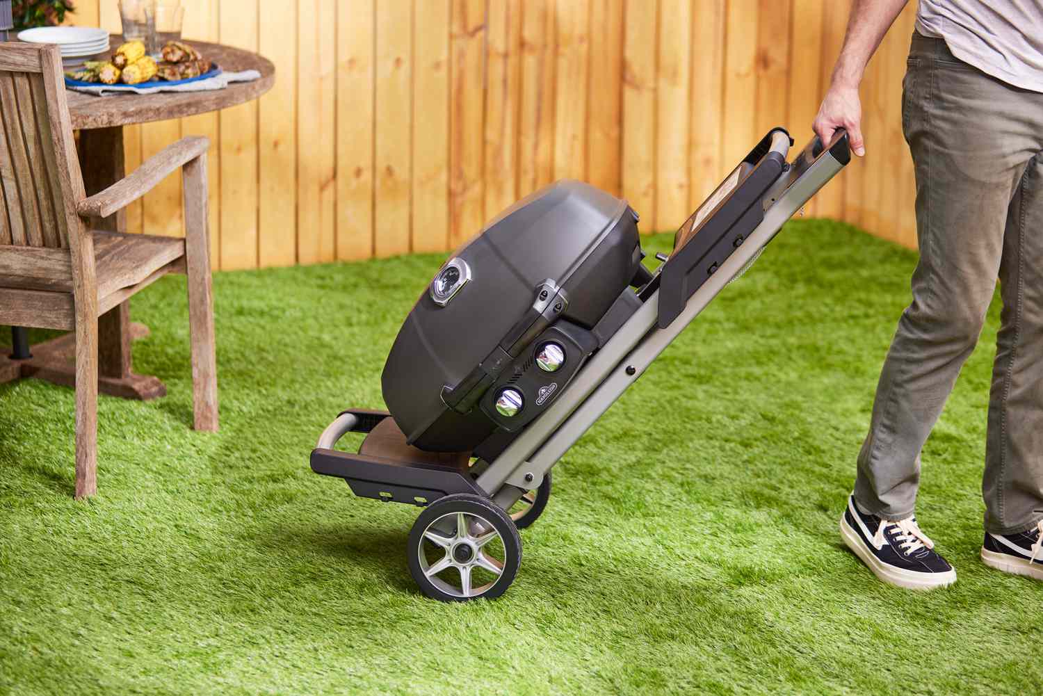 A person rolling a portable gas grill across a grass surface.