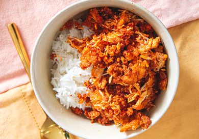 A bowl of white rice topped with a spiced chicken dish, served alongside a gold utensil on a cloth background