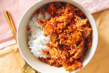 A bowl of white rice topped with a spiced chicken dish, served alongside a gold utensil on a cloth background