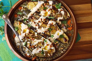 Overhead shot of a bowl of stewed lentils topped with a halved, roasted eggplant, drizzled with tahini sauce and sprinkled with toasted pine nuts.