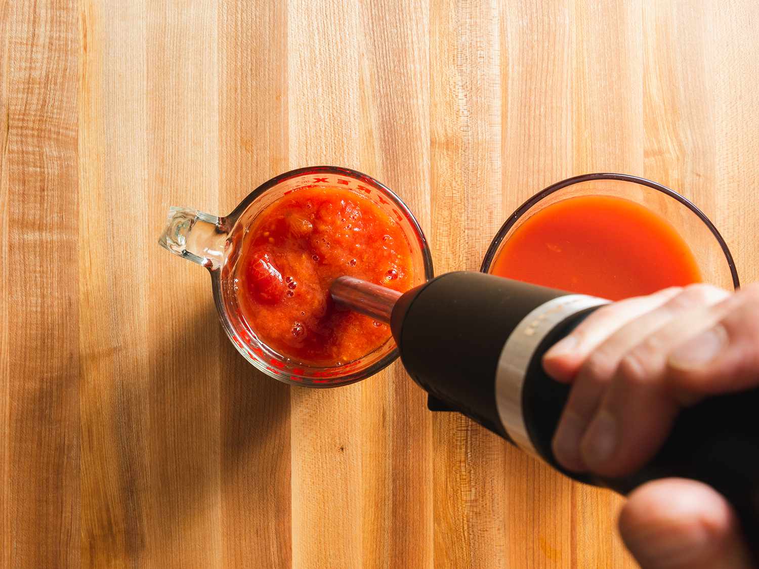 Whole canned tomatoes being blended with an immersion blender in a measuring cup with reserved tomato liquid alongside