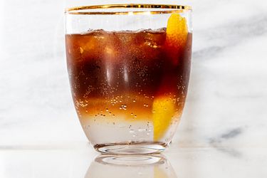 A clear glass filled with a layered drink of espresso on the top and tonic water on the bottom, with a stip of lemon peel in the glass and some ice.
