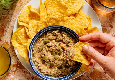 A hand dipping a tortilla chip into a bowl of dip garnished with seeds surrounded by tortilla chips on a plate