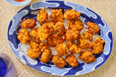 Fritters on a blue and white printed plate, on a bamboo mat. Glass of blue water, and red chili sauce on the side 