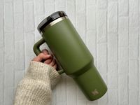 A hand holding a green Stanley Quencher ProTour tumbler in front of a white brick background