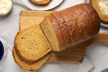 Sliced loaf of anadama bread on a wooden board, with two slices on plates, with grey striped napkin, mug of coffee, plate, butter, and breadknife behind it 