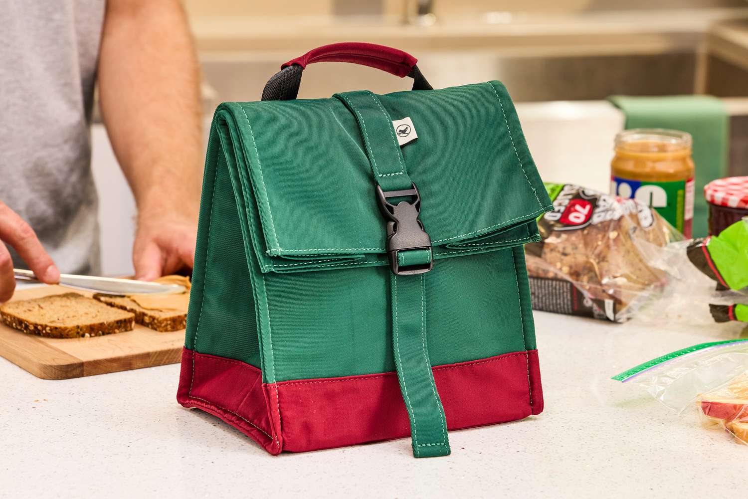 A green and red lunchbox in front of a person making a peanut butter sandwich