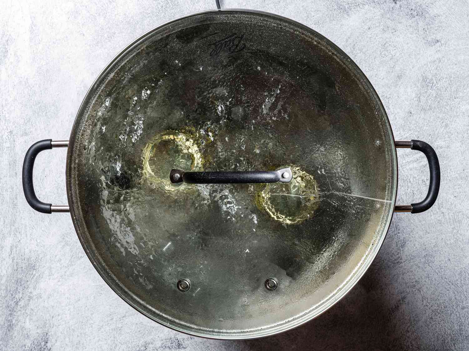 A top-down shot of a stainless steel pot of boiling water covered by a glass lid. Inside of the water are the two jars of Brussels sprouts, which are being processed so that they are shelf stable.