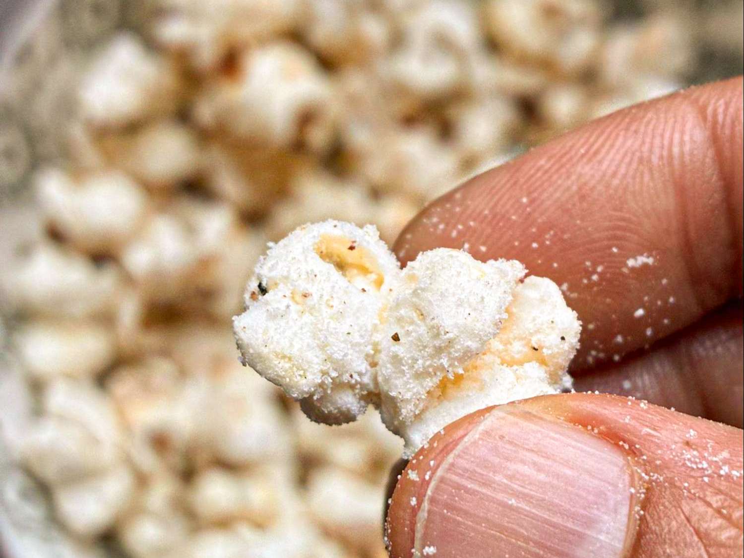 A closeup of a hand holding a single piece of salted popcorn with a bowl of popcorn in the background