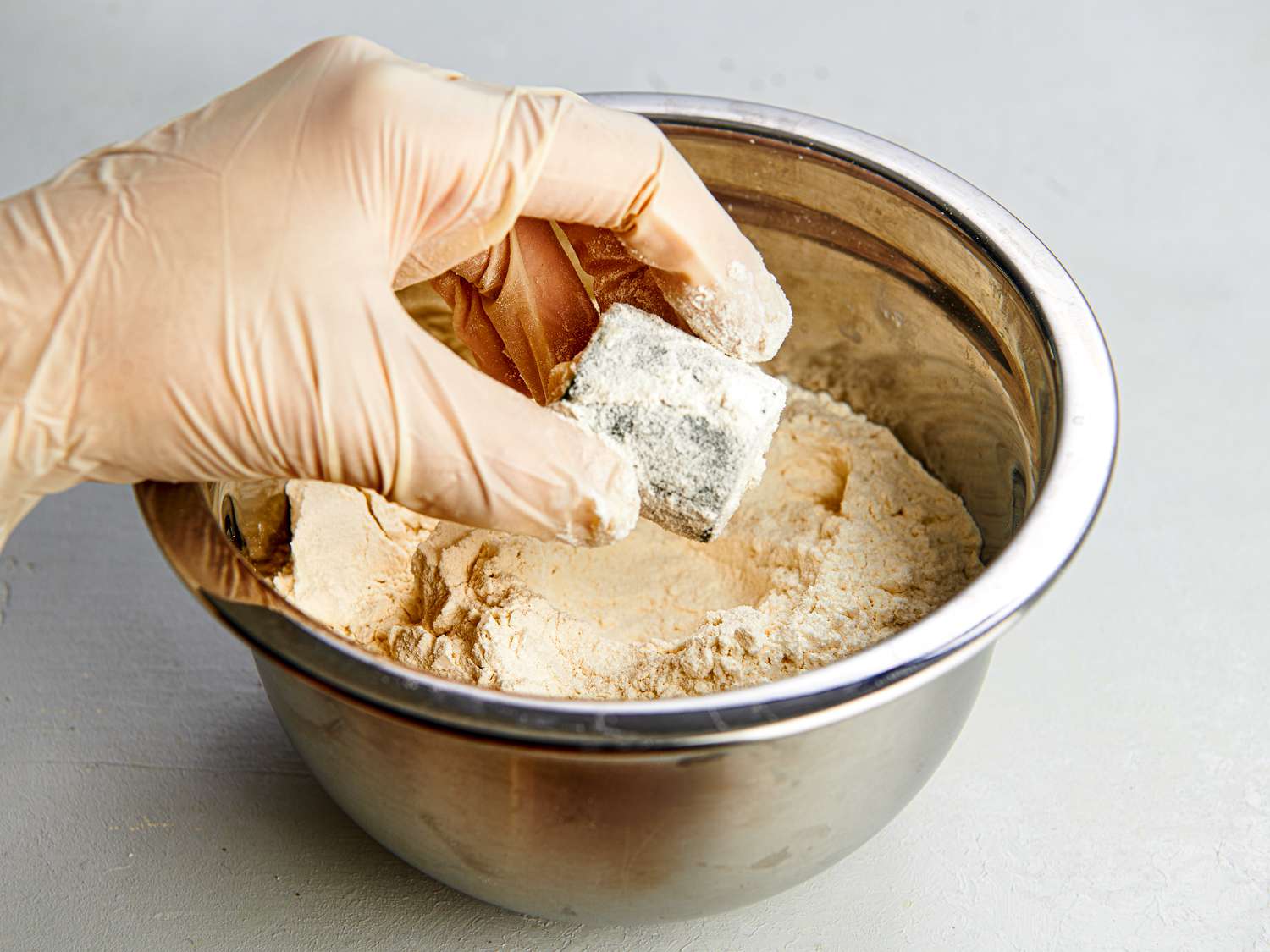 A gloved hand holding a stuffed jalapeÃ±o ring, which had been dipped in milk and now is being removed from a metal mixing bowl holding flour. The image shows the ring evenly coated in flour.