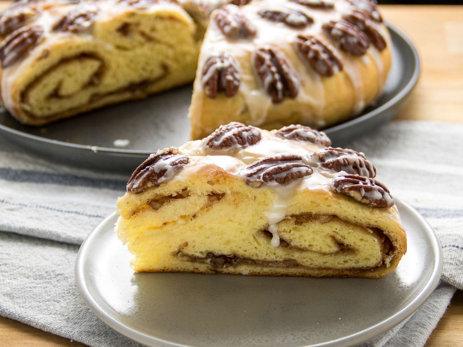 slice of yeasted coffee cake on a plate with full cake in the background, on a wooden tabletop and grey napkins