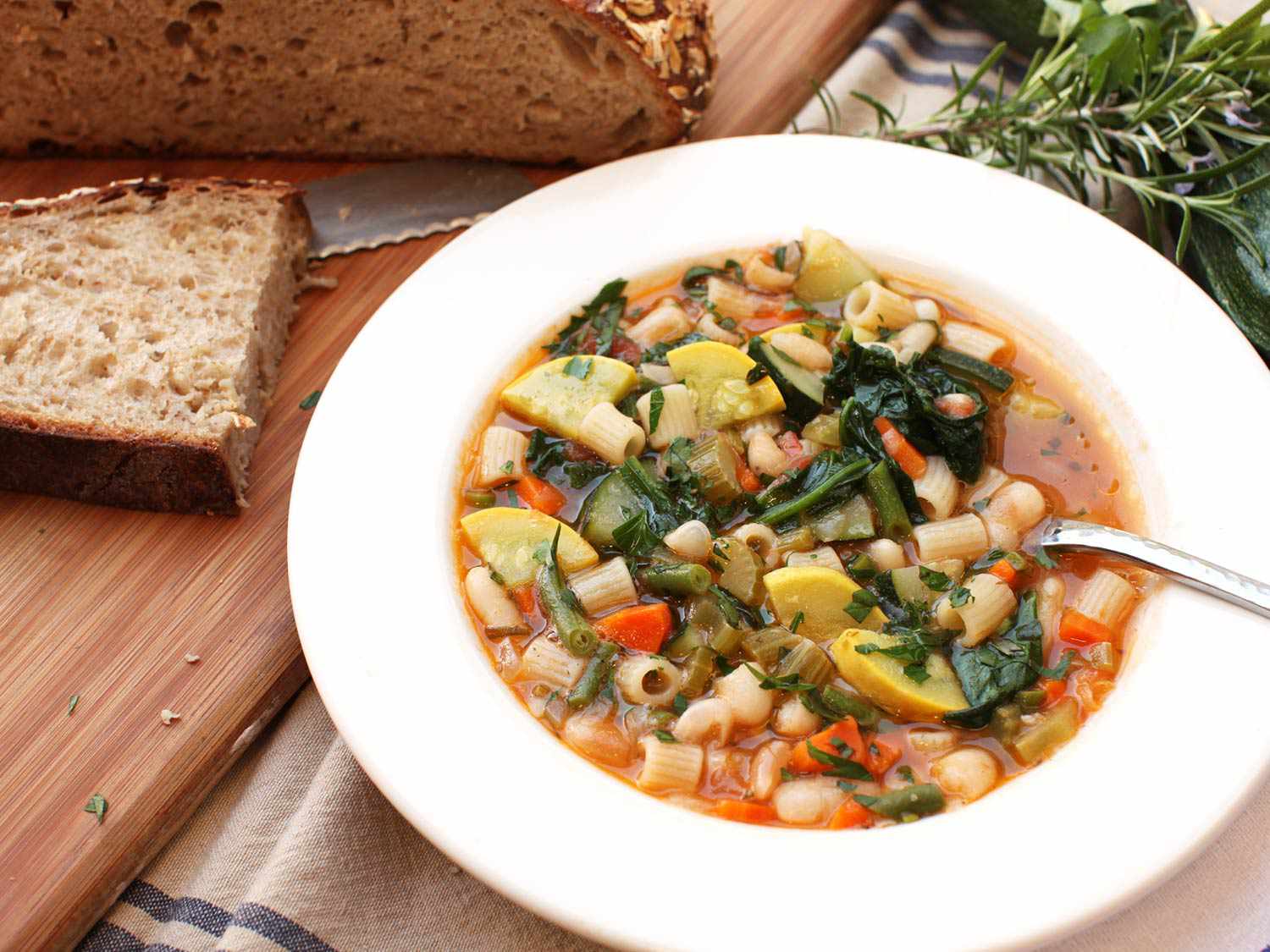 A bowl of minestrone soup in a white bowl with a sliced loaf of bread off to the side.