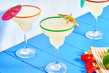 Three margaritas on a blue tile surface, each garnished with small paper umbrellas, with a decorated napkin and lime nearby