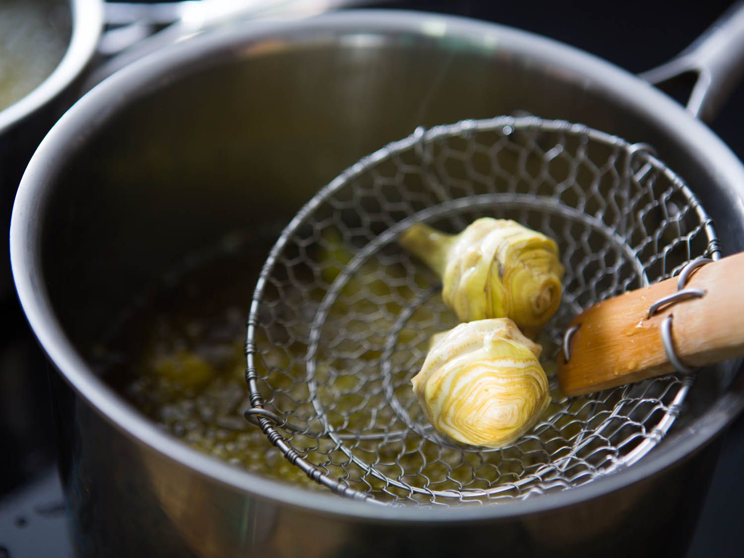 A couple of trimmed artichoke hearts in a spider being lowered into hot oil.