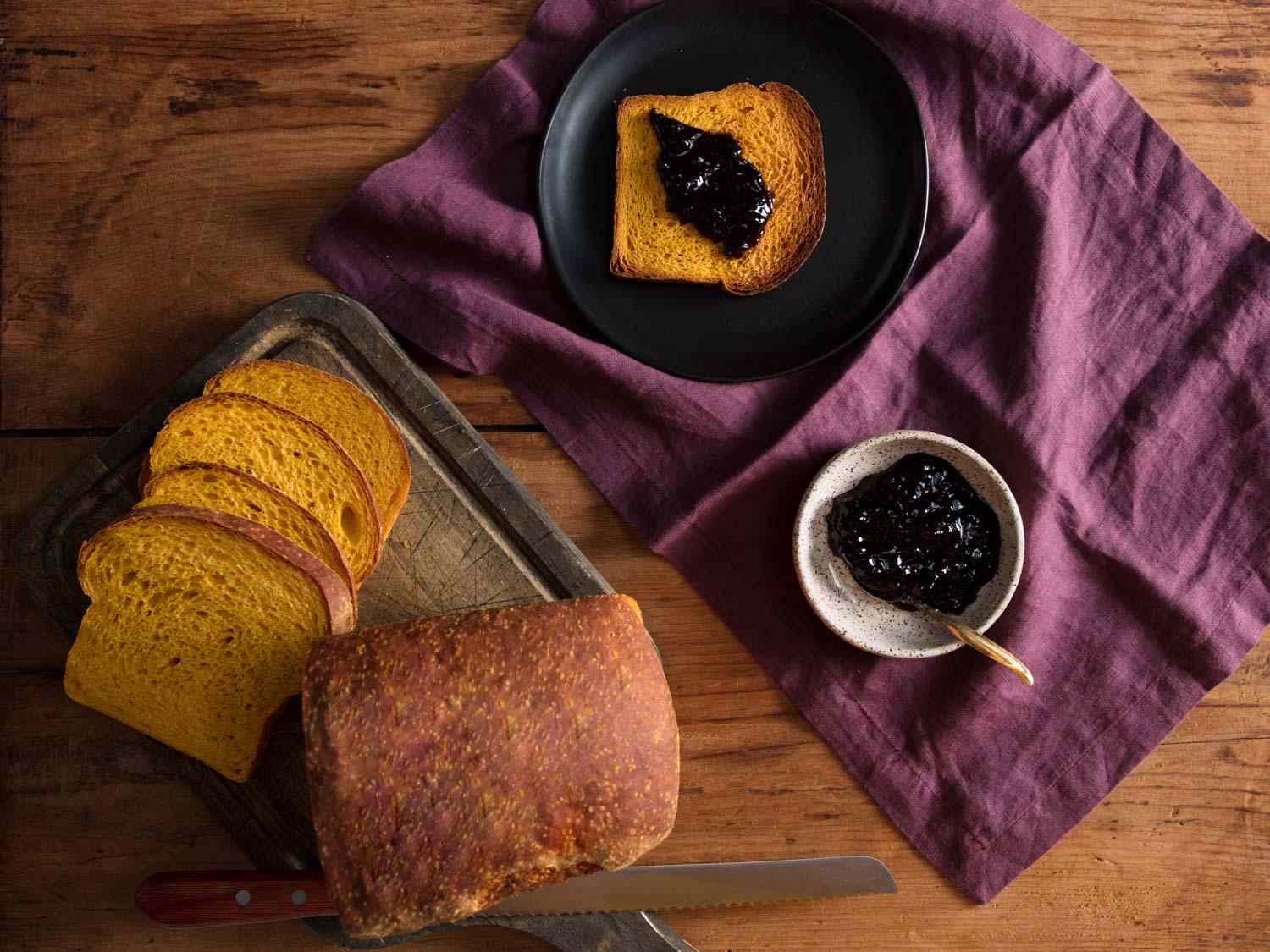 A loaf of pumpkin sandwich bread sliced into five slices, with the rest of the loaf intact. One slice is spread with dark jam and the jam bowl is on the side.