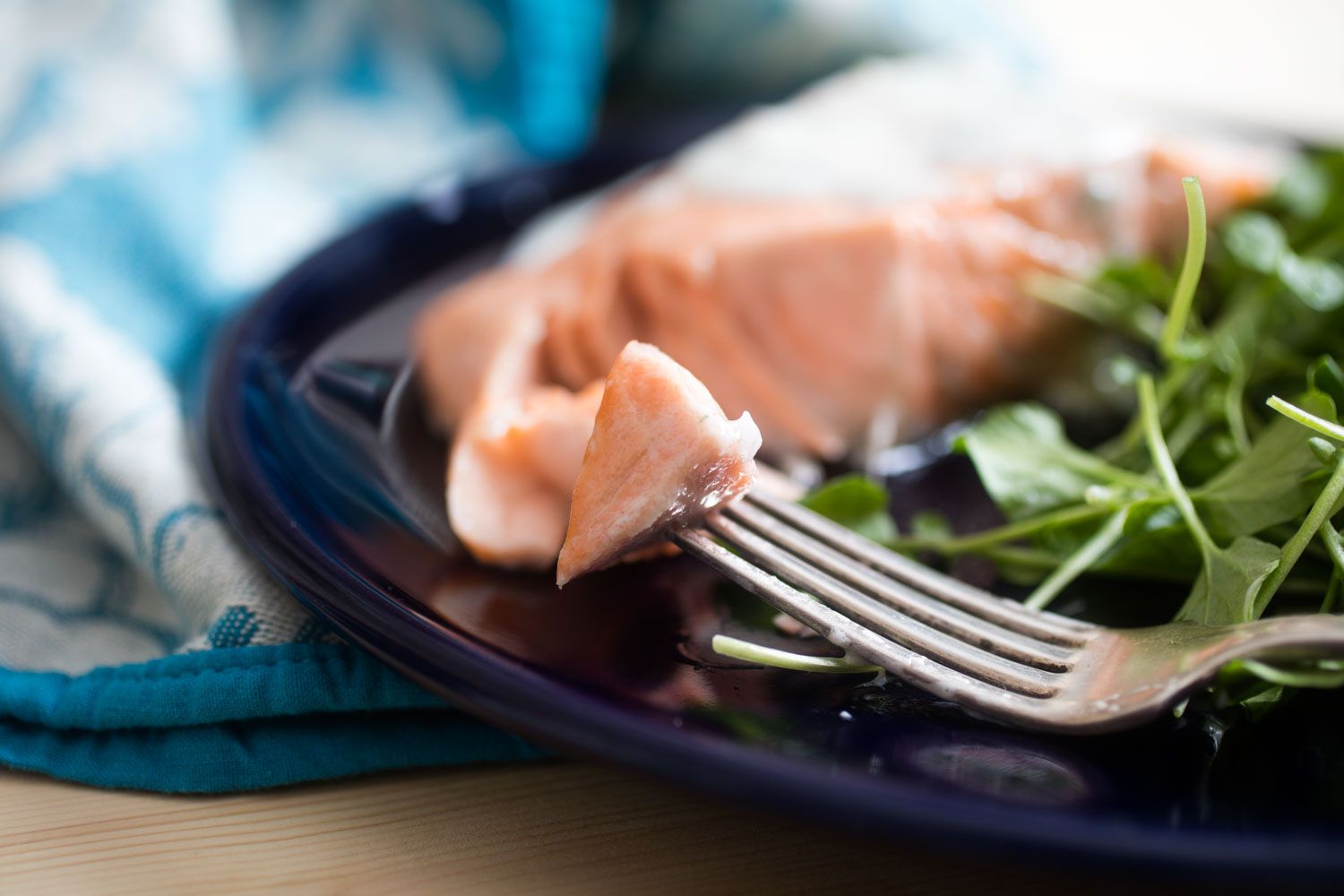 A bite of salmon skewered on the tines of a fork. The fork is resting on a dark blue plate. More salmon and dressed greens are in the blurred background. 