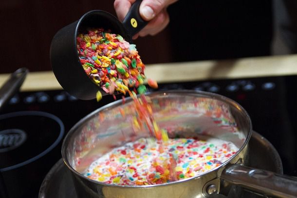 Pouring a cup of fruit pebble cereal into ice cream base in a pan on the oven.