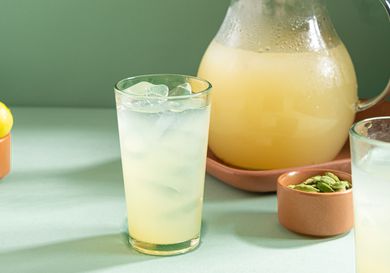 A glass of lemonade with ice cubes beside a pitcher containing more lemonade, next to small bowls of cardamom and lemons