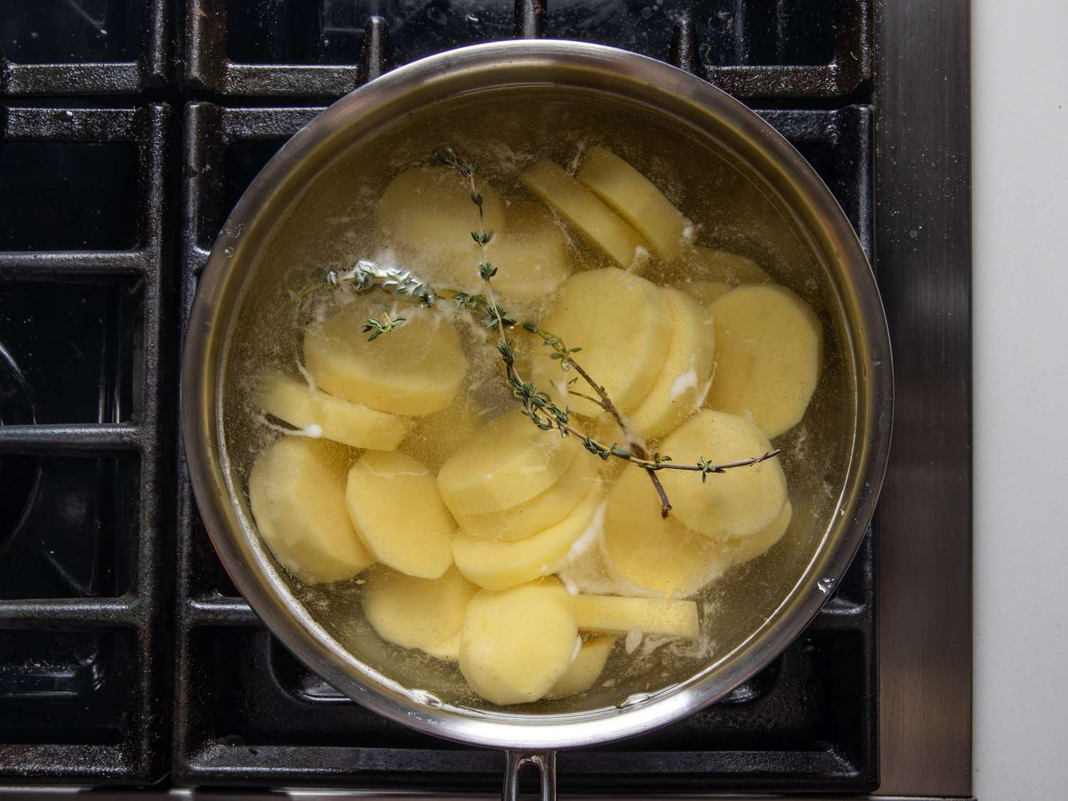 Sliced Yukon Gold potatoes sit in a pot of water with a couple thyme sprigs.