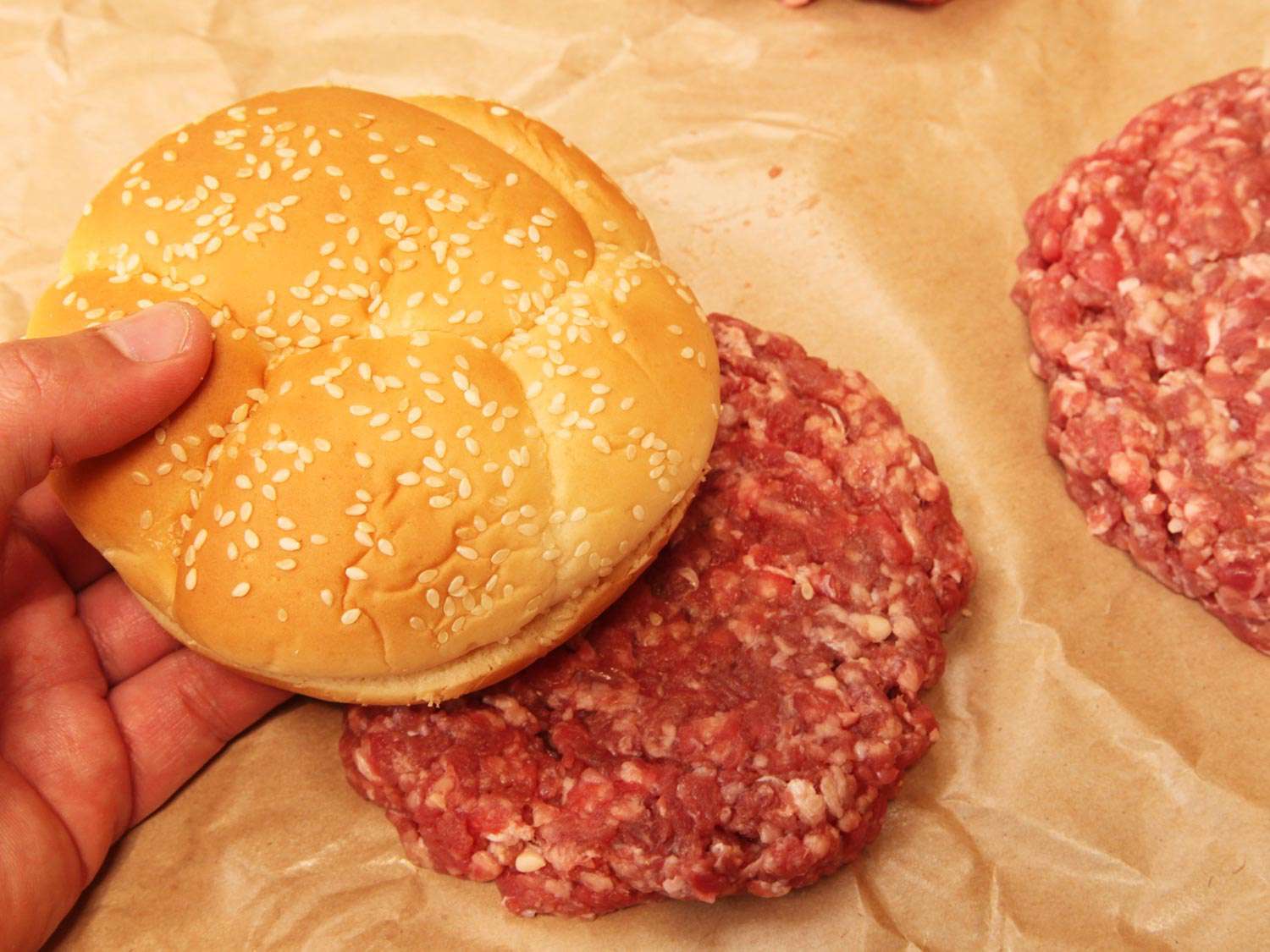 Holding a sesame bun up to a raw burger patty to show that the patty is slightly bigger than the bun.