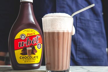 A glass of chocolate egg cream with a metal straw next to a bottle of Ubet chocolate syrup