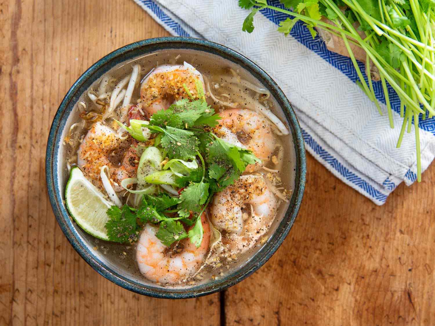 Overhead shot of kuy teav phnom penh (Cambodian soup with rice noodles), with poached shrimp, cilantro leaves, lime wedge, scallions, and bean sprouts